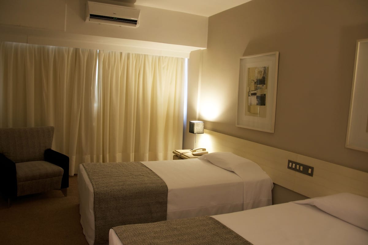 A hotel room with two beds, a chair, a lamp, and a painting on the wall.