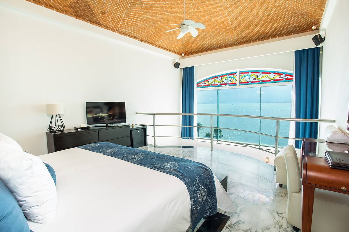 A modern hotel room with a large bed, a desk, a television, and a balcony with a view of the sea.