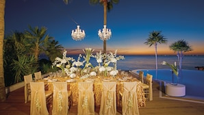 An outdoor dining area with a table set for a meal, surrounded by chairs with golden sashes, and two chandeliers above.