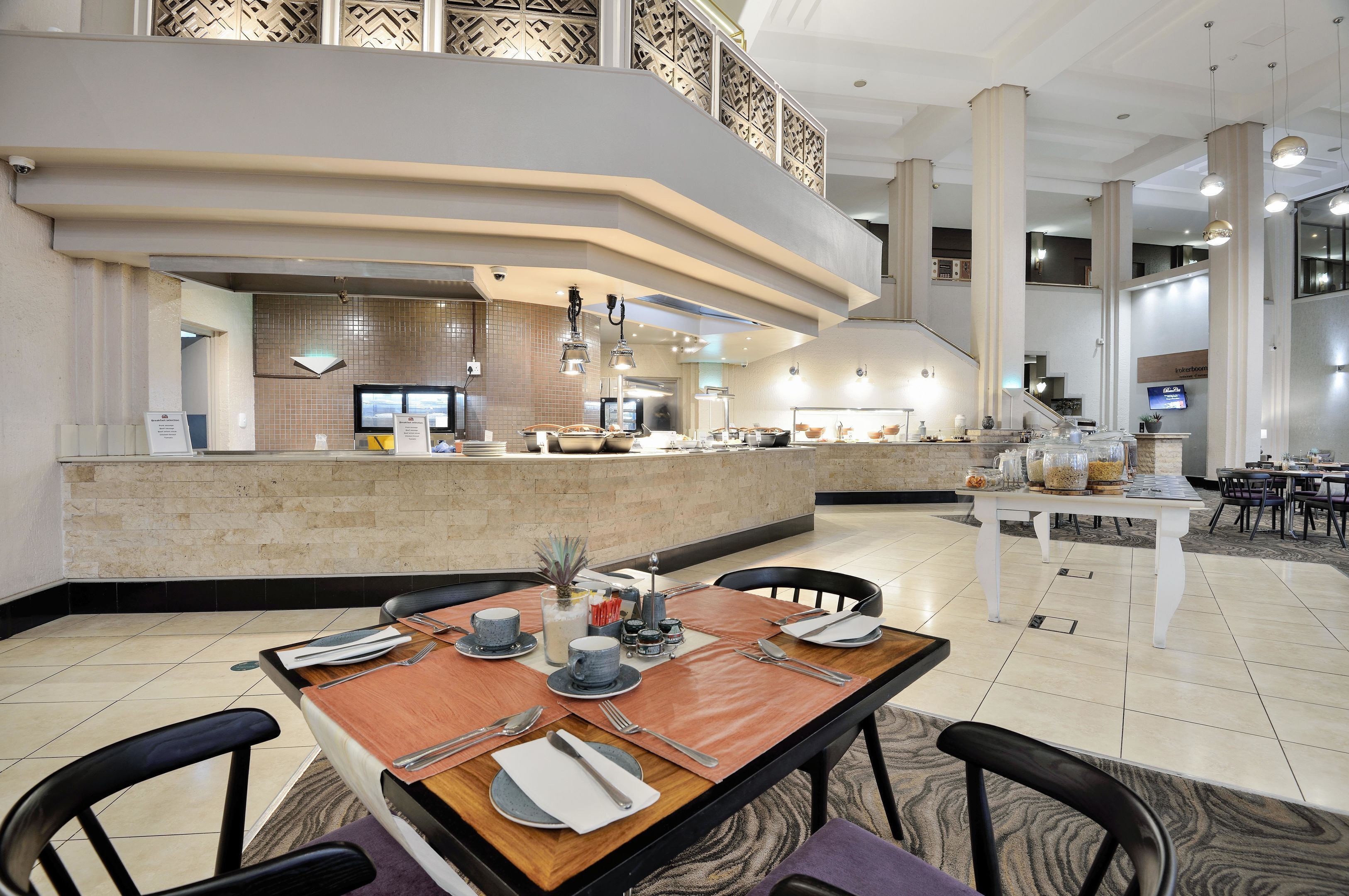 A modern hotel lobby with a central dining area, a reception desk, and a staircase leading to an upper level.