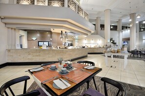 A modern hotel lobby with a central dining area, a reception desk, and a staircase leading to an upper level. - Windhoek Country Club (Windhoek)