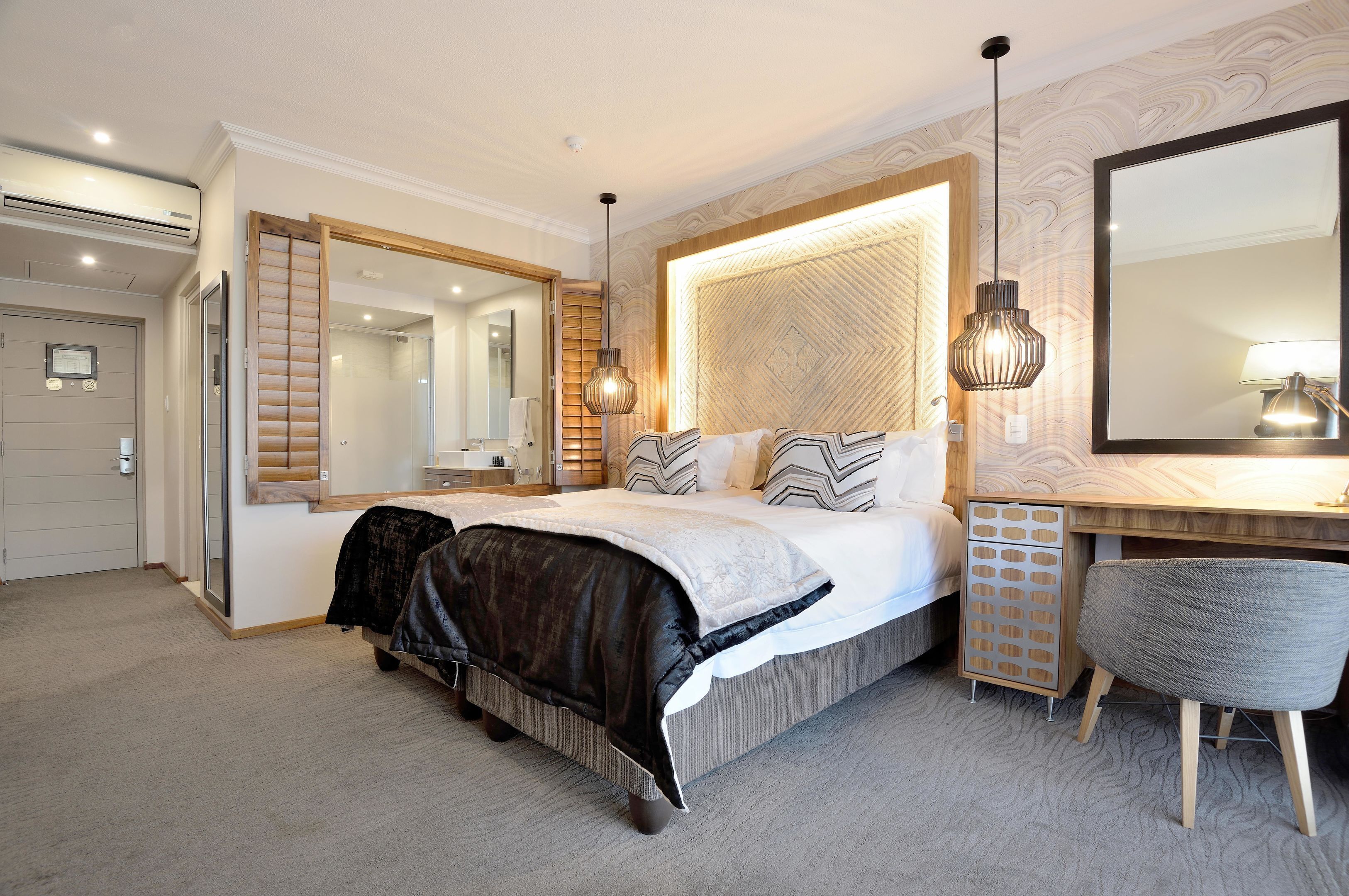 A modern hotel room with a large bed, a desk, a chair, a mirror, and a built-in wardrobe.