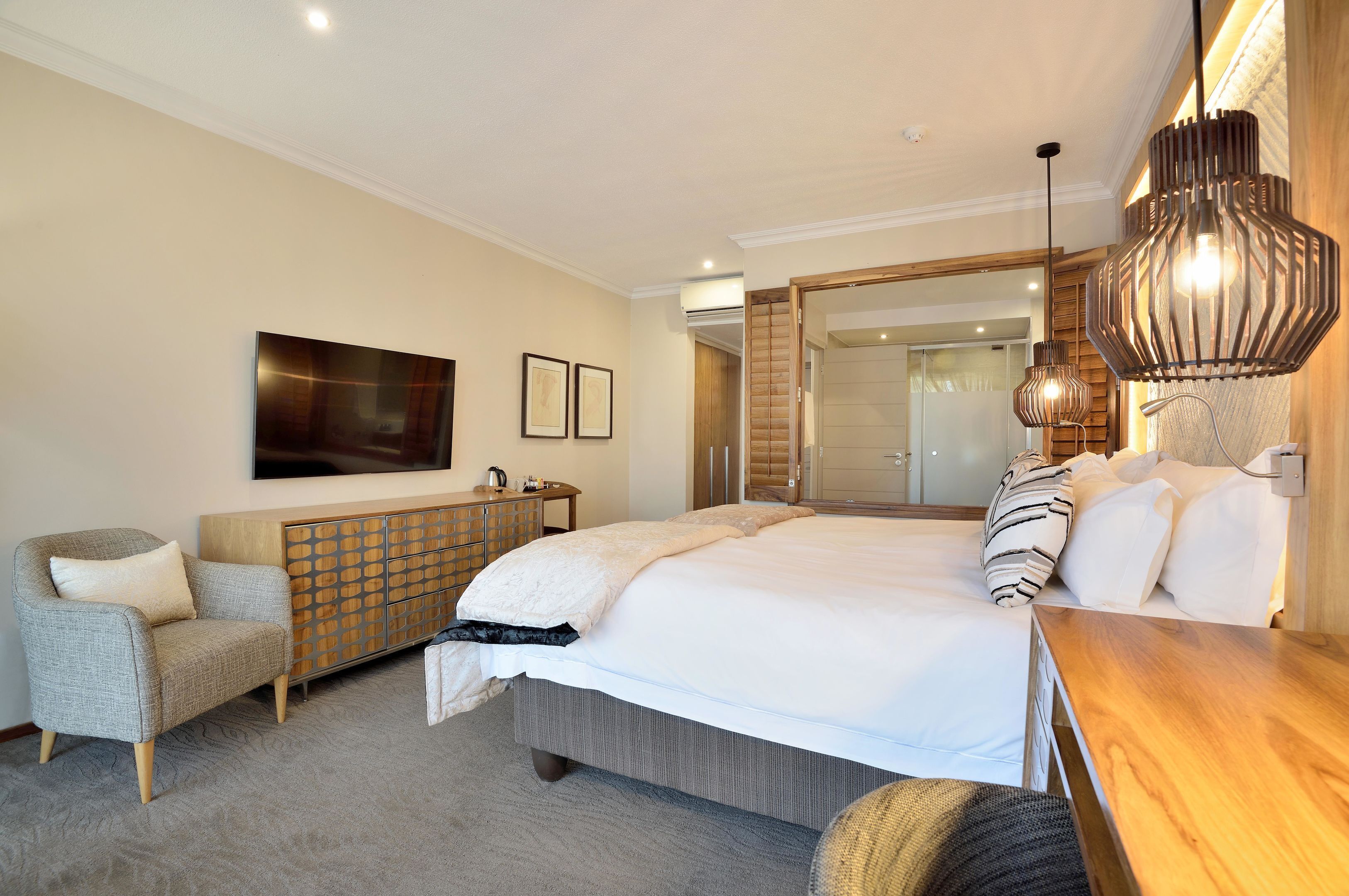 A modern hotel room with a large bed, a wooden side table, a seating area with a chair, a large mirror, and a television mounted on the wall.