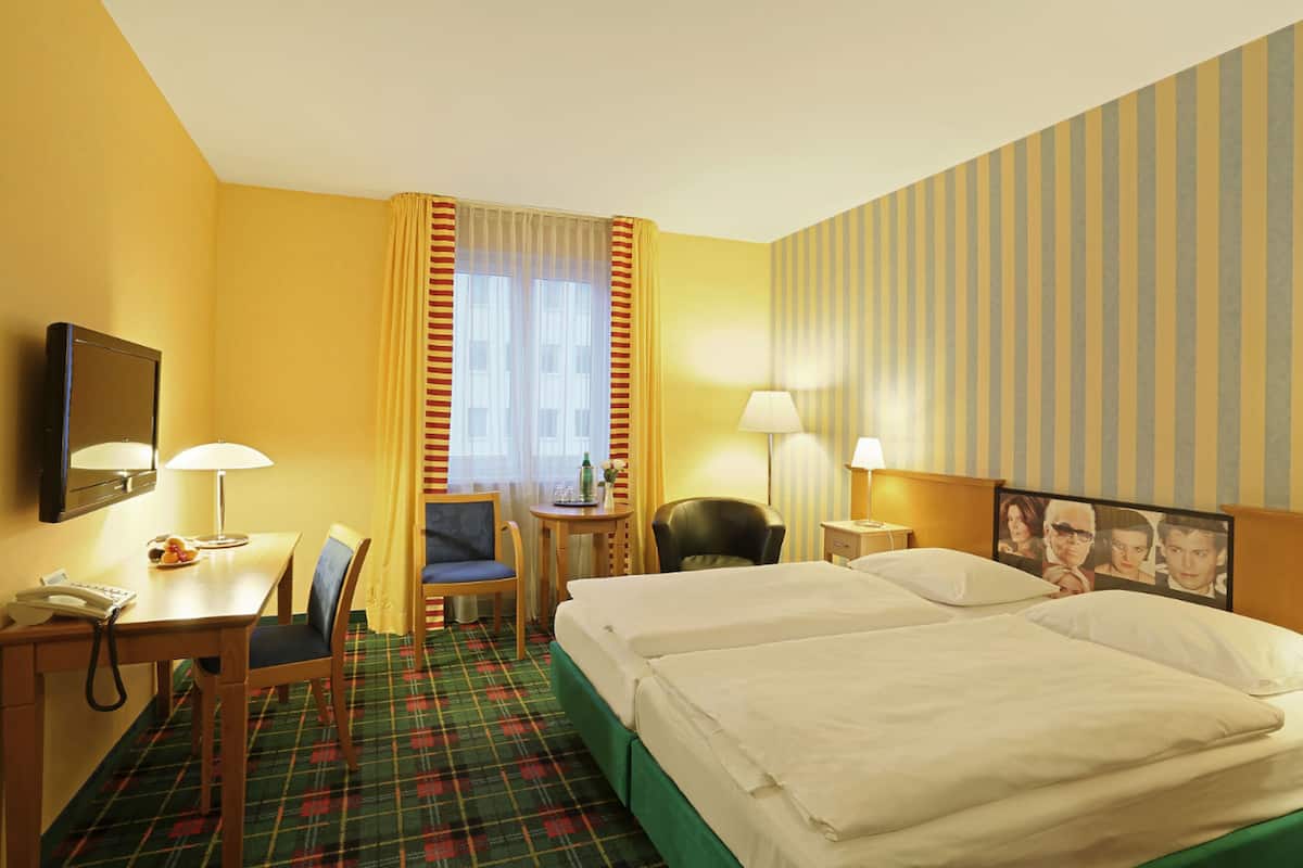 A hotel room with two beds, a desk with a telephone, a chair, a small table with a lamp, and a television on the wall.