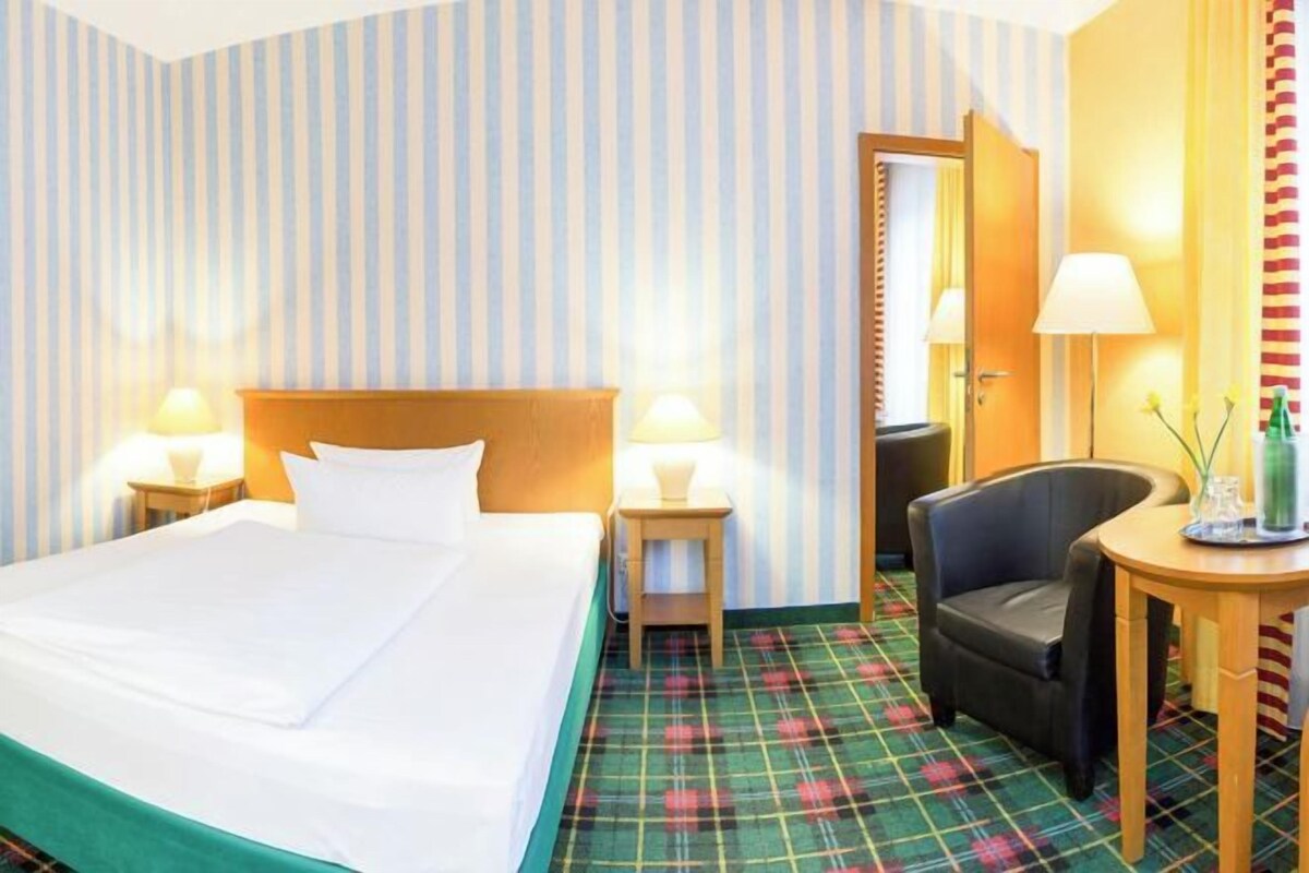 A hotel room with a bed, a chair, a small table, and a lamp.