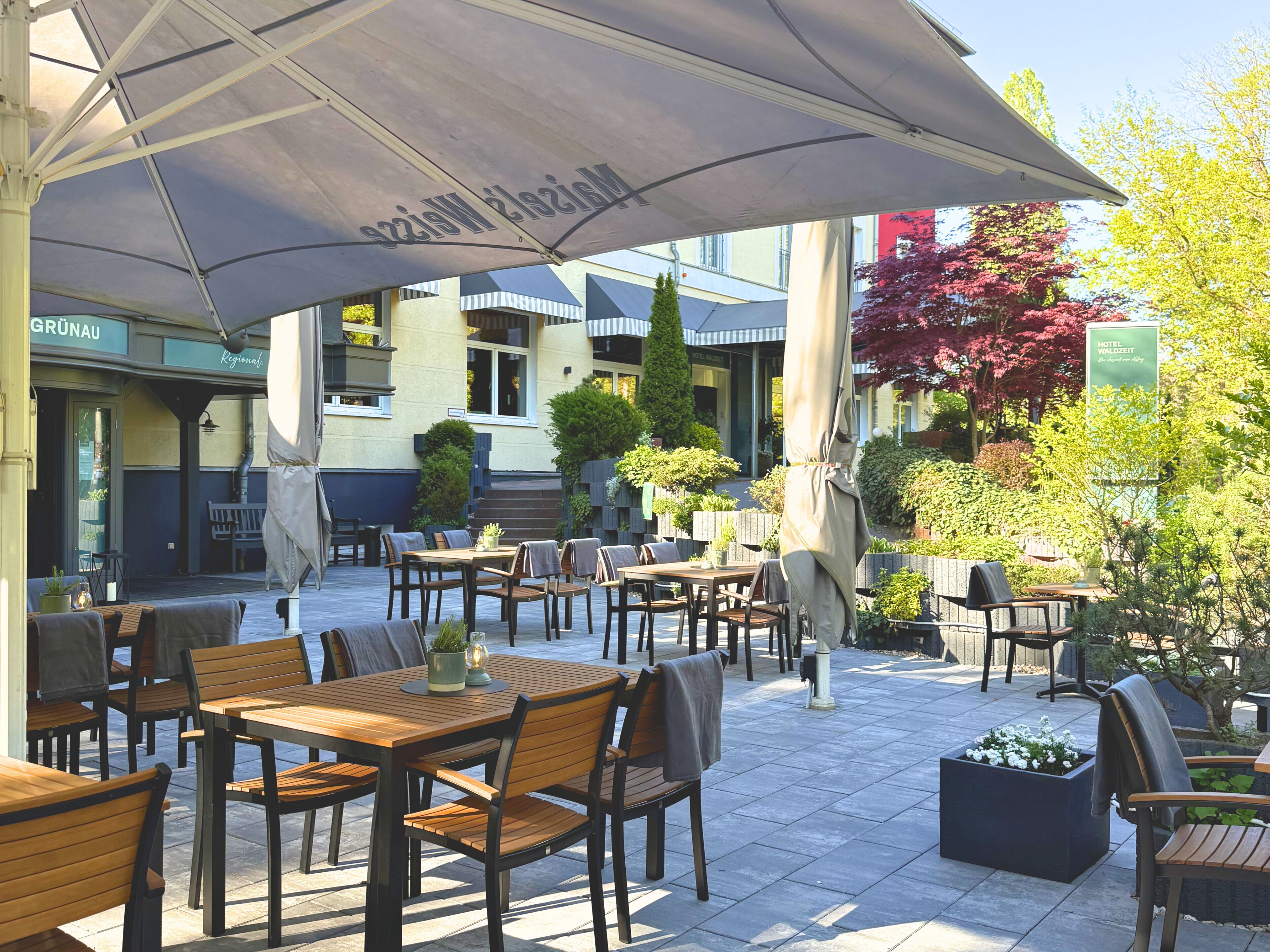 Een terras met tafels en stoelen, overdekt door een grote parasol, omgeven door groen en gebouwen.