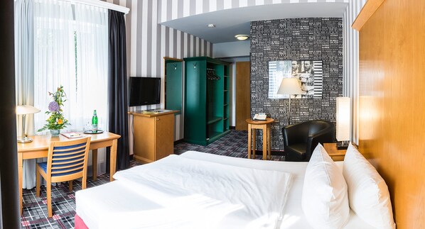 A hotel room with a bed, a desk with a chair, a TV, a wardrobe, a small table with a lamp, and a wall with a quote mural. - Hotel Waldzeit Berlin - Grünau (Berlin)