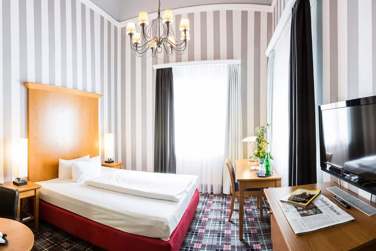 A hotel room with a bed, a desk with a TV, a chair, a lamp, and a window with curtains.