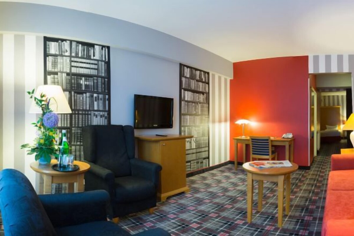 A hotel room with a black armchair, a wooden desk, a television, and a bookshelf.