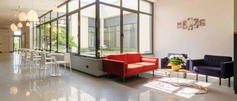 Un vestíbulo amplio con diseño moderno, que incluye un sofá rojo, un sillón morado y una mesa de centro con un jarrón de flores. Amplias ventanas dejan entrar luz natural al espacio.
