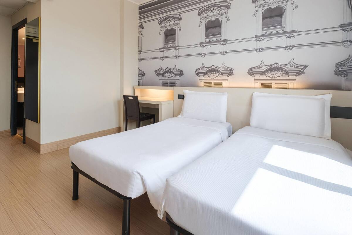 A hotel room with two single beds, a desk with a chair, and a wall with a patterned design.