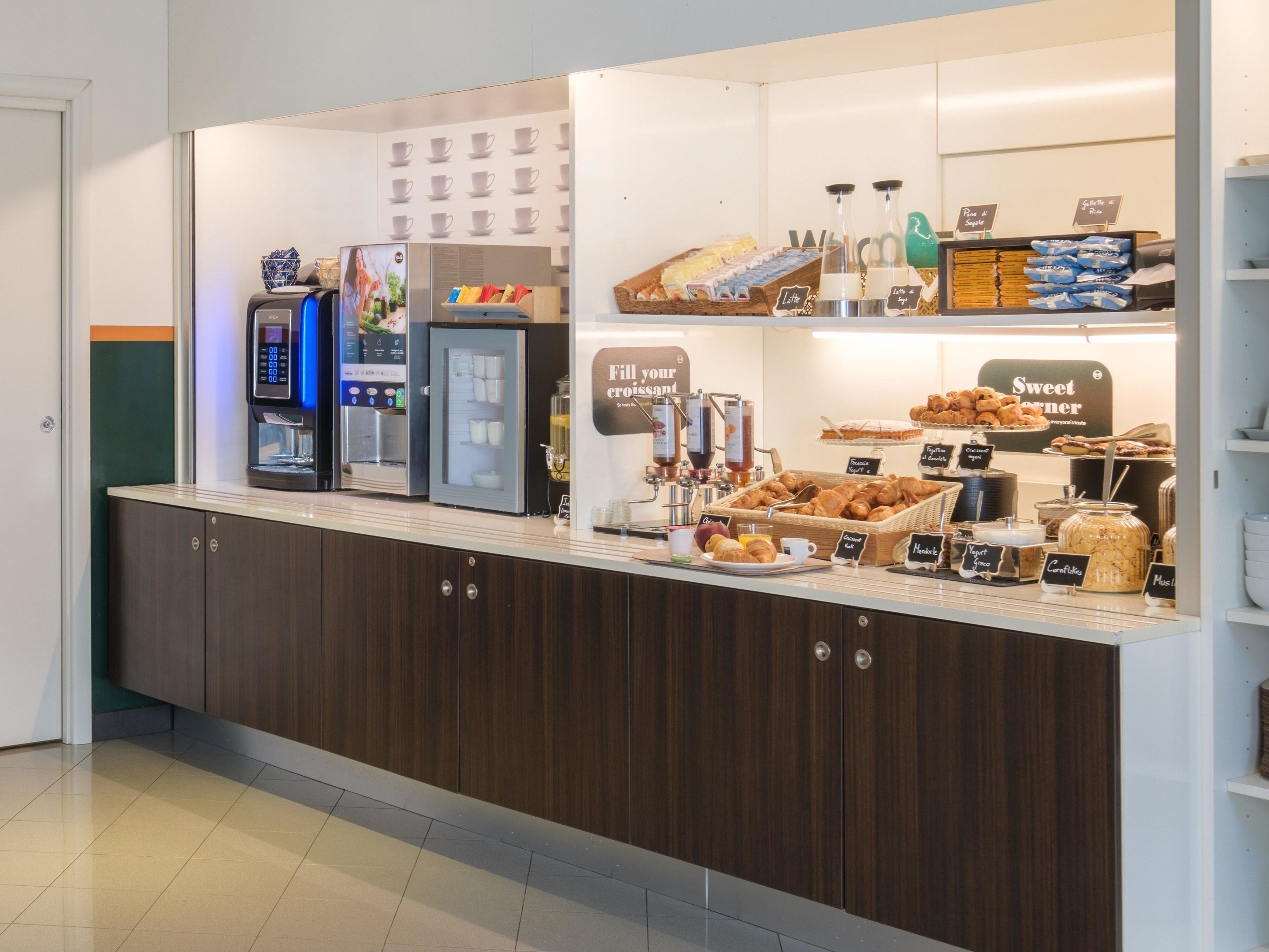 A self-service coffee station with espresso machines, a refrigerated display case, and a variety of baked goods.