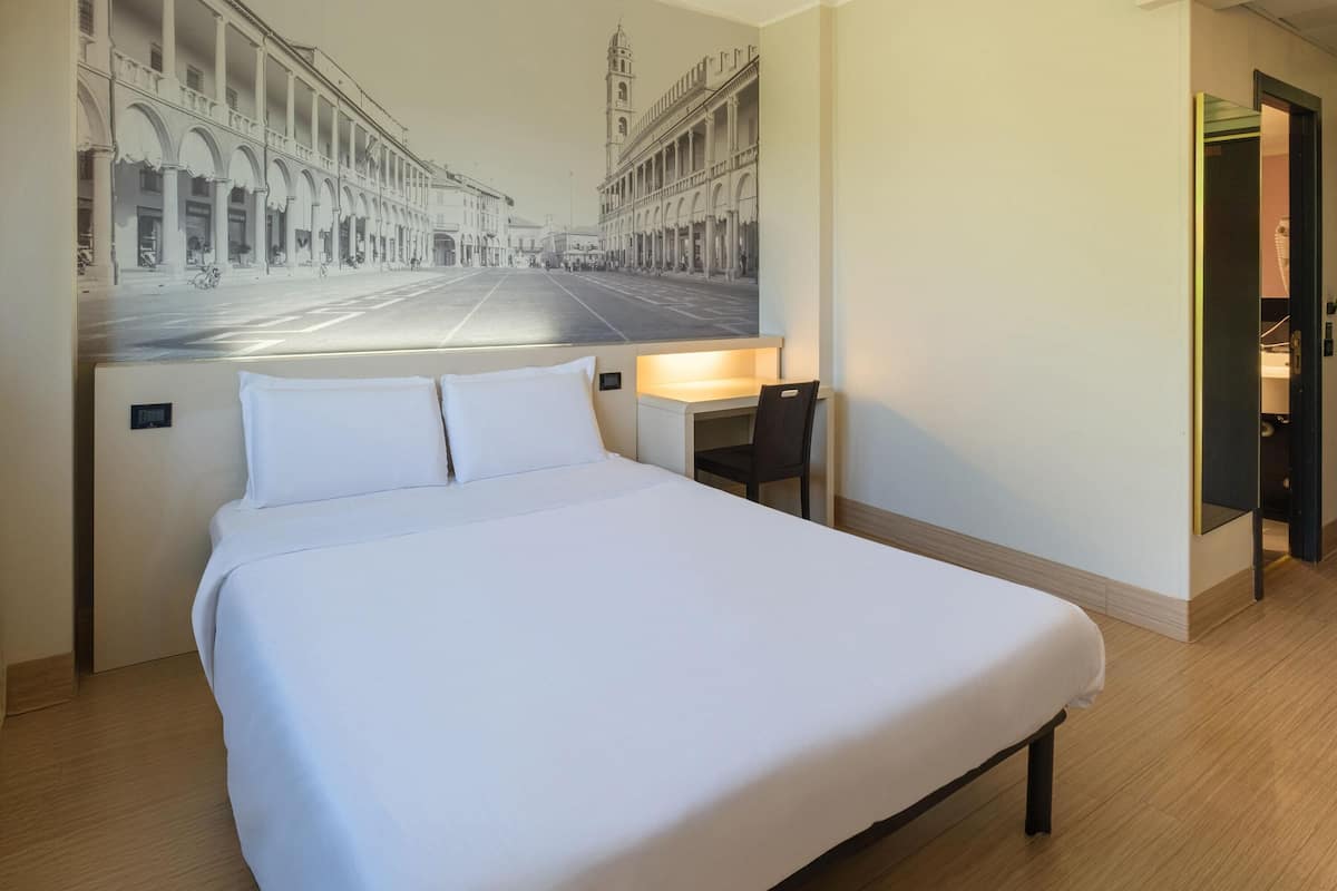 A hotel room with a large bed, a desk, and a chair. There is a wall mural of a city street.