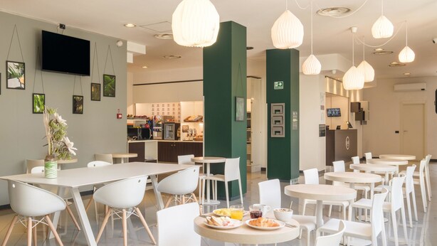 A modern dining area with white tables and chairs, a kitchen in the background, and pendant lighting. - B&B Hotel Faenza (Faenza)