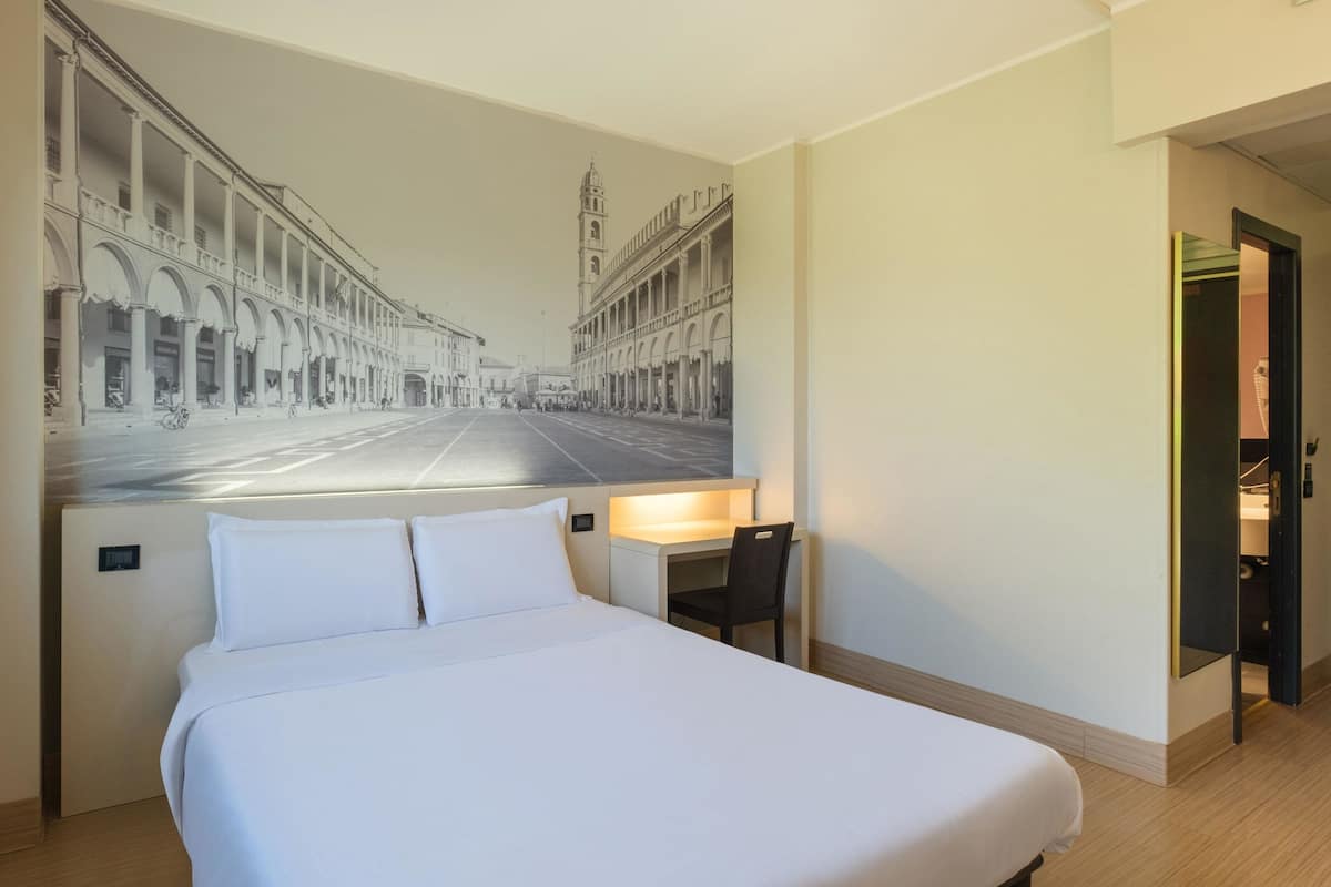 A hotel room with a large bed, a desk, and a chair. There is a mural on the wall depicting a street scene.