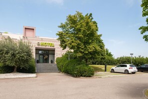 A hotel with a sign reading "HOTEL" in green letters, a paved entrance, and a parking area with a white car. - B&B Hotel Faenza (Faenza)