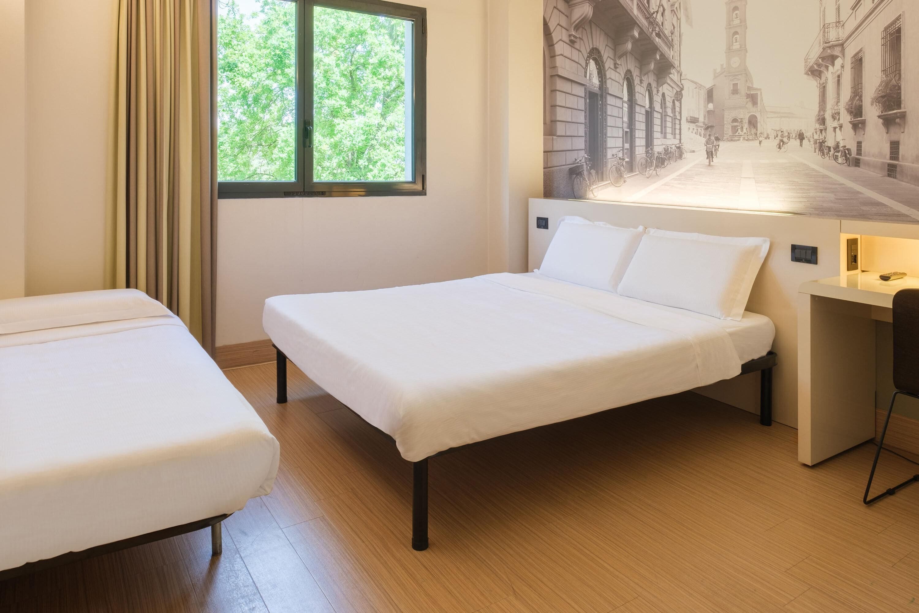 Een hotelkamer met twee enkele bedden, een bureau, een stoel en een groot raam met gordijnen.