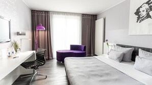 A modern hotel room with a bed, a desk, a chair, a purple ottoman, a desk lamp, and a painting on the wall. - elaya hotel wolfenbuettel, Trademark Collection by Wyndham (Wolfenbuettel)