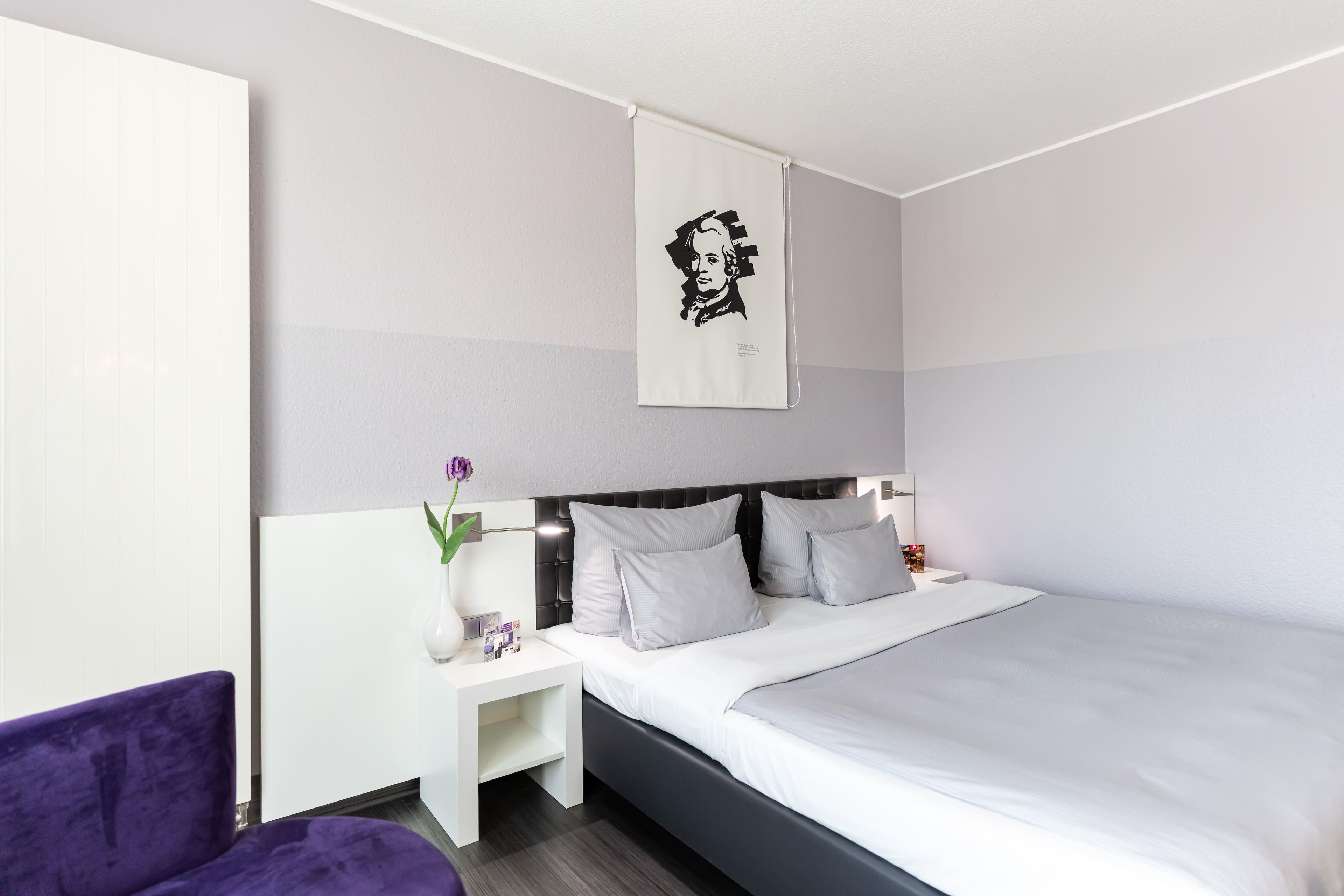 Una habitación de hotel moderna con una cama grande, una mesita de noche, una silla morada y una obra de arte colgada en la pared.