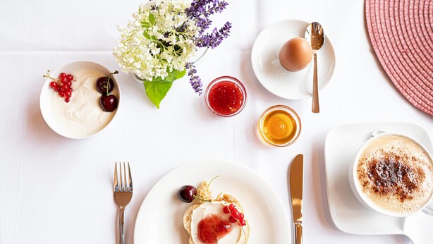Un desayuno con tostadas cubiertas de crema y frutos rojos, una taza de café con canela y un tazón de yogur con cerezas.