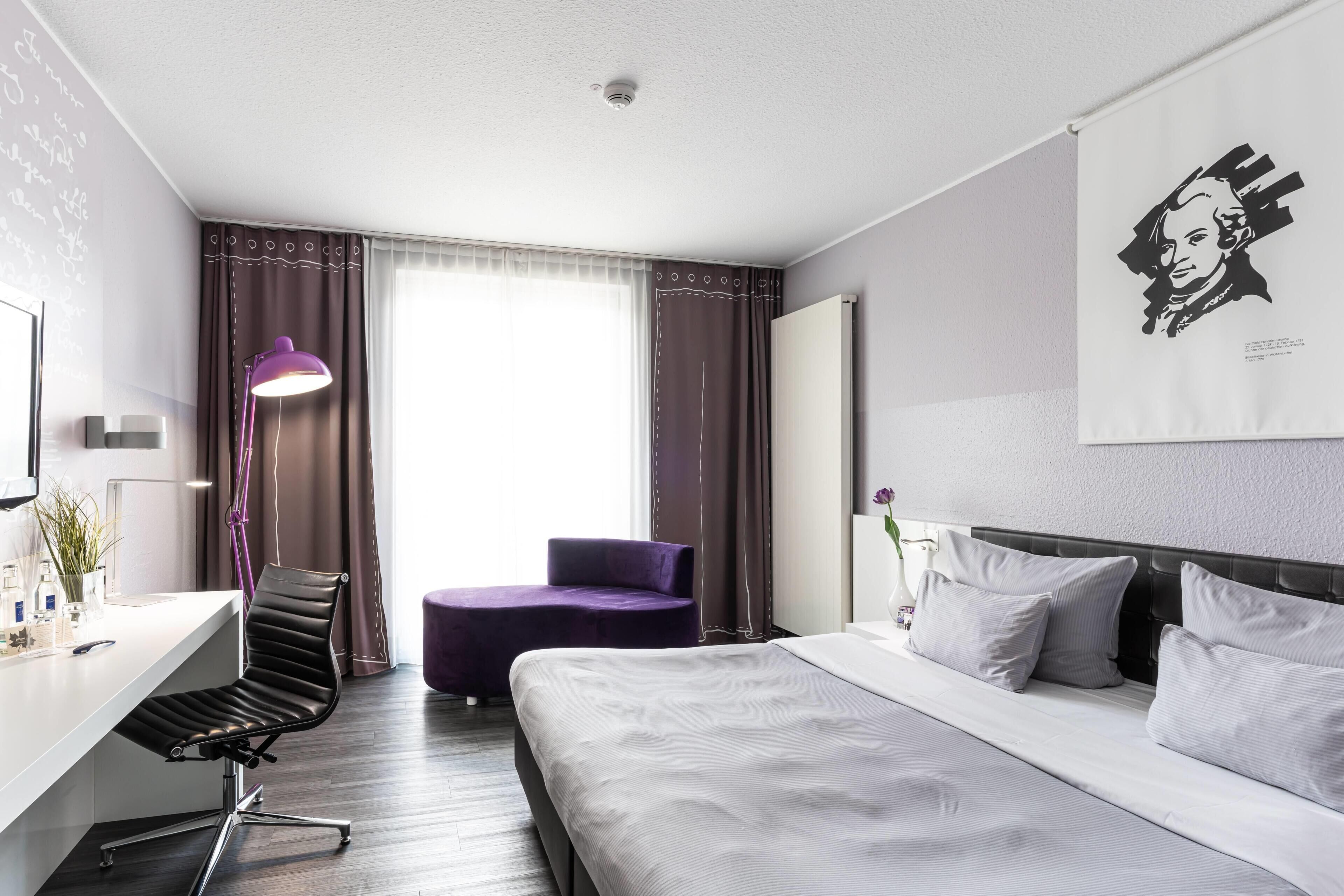 Une chambre d’hôtel avec un lit, un bureau, une chaise, une lampe et une fenêtre avec des rideaux.
