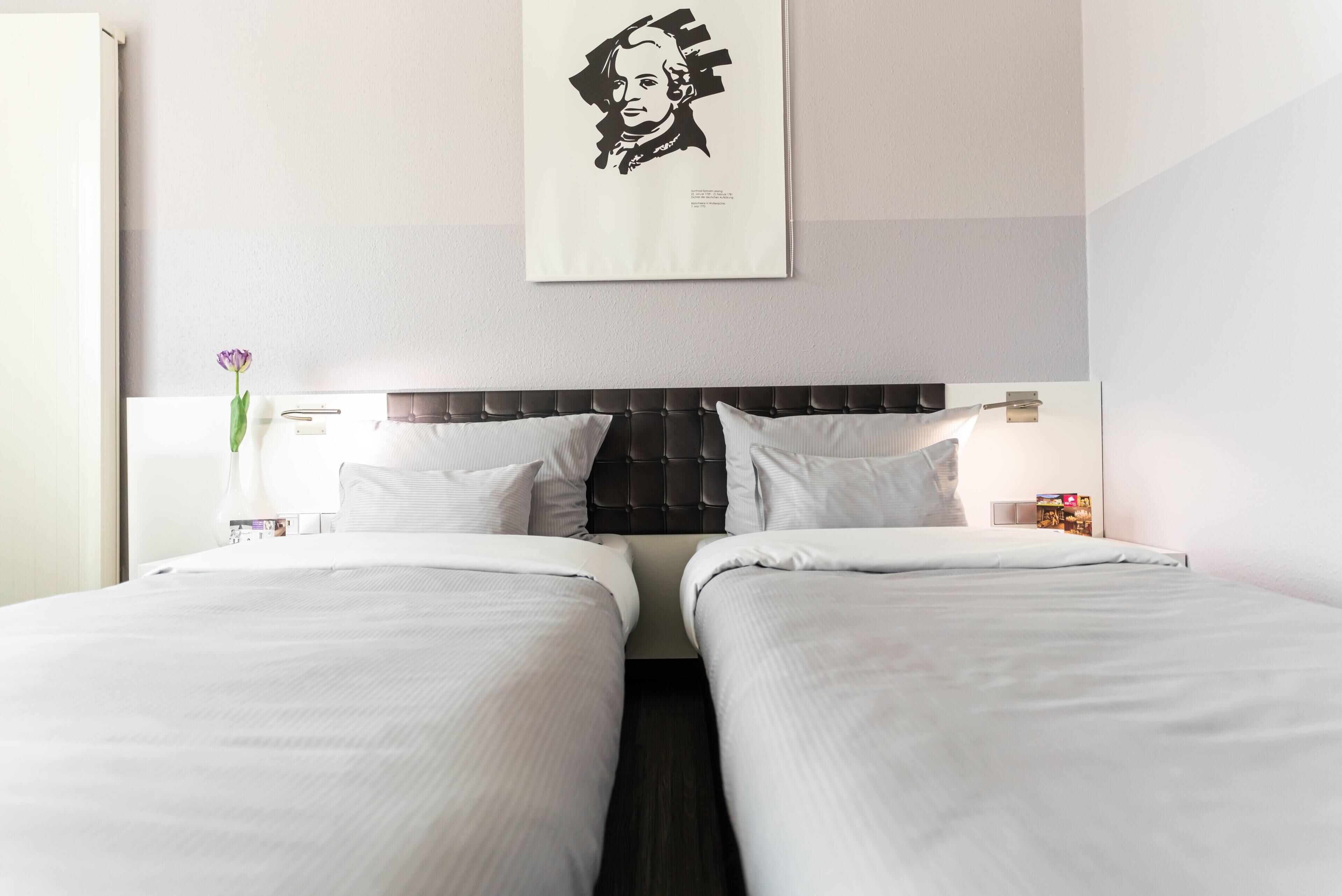 A hotel room with two beds, a dark headboard, white bedding, a framed portrait on the wall, a vase with a flower, and a nightstand with a lamp.