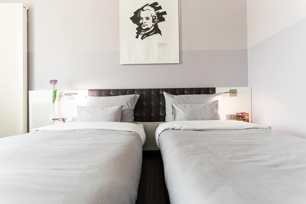 A hotel room with two beds, a dark headboard, white bedding, a framed portrait on the wall, a vase with a flower, and a nightstand with a lamp.