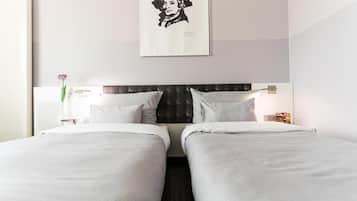 A hotel room with two beds, a dark headboard, white bedding, a framed portrait on the wall, a vase with a flower, and a nightstand with a lamp.