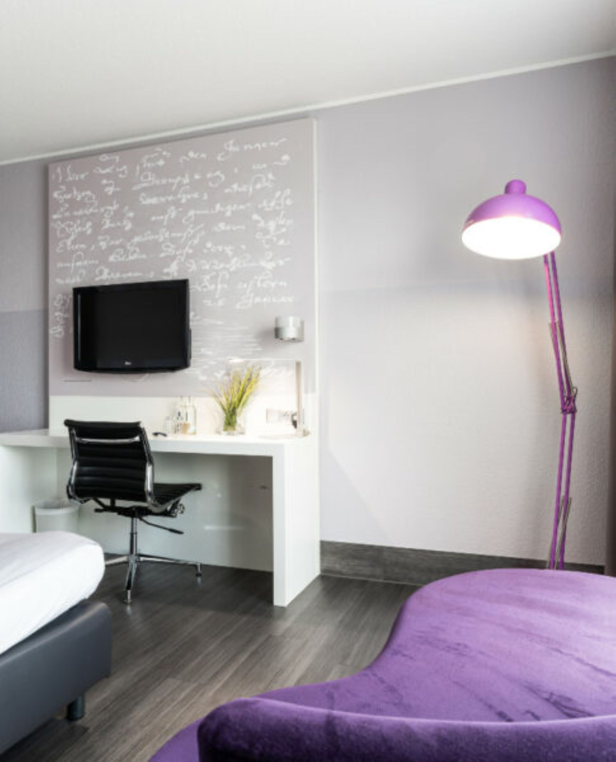 Una habitación de hotel moderna con televisión de pantalla plana montada en la pared, un escritorio con silla, una mesita de noche y una colcha púrpura.