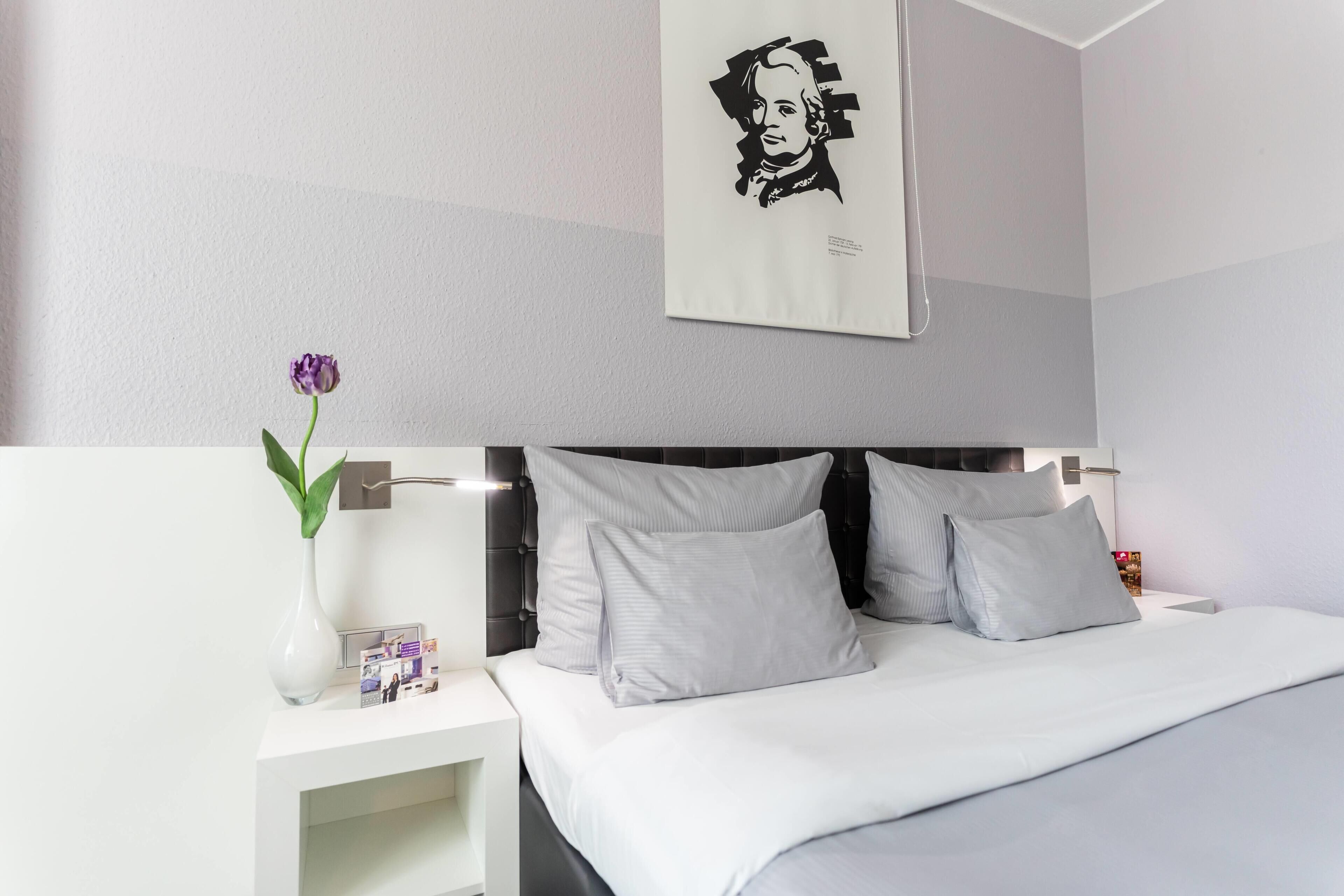 A modern hotel room with a bed, white nightstands, a vase with a flower, and a wall-mounted picture.