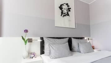 A modern hotel room with a bed, white nightstands, a vase with a flower, and a wall-mounted picture.