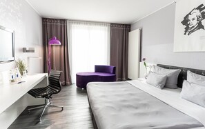 A hotel room with a bed, a desk, a chair, a purple ottoman, a lamp, and a painting on the wall. - elaya hotel wolfenbuettel, Trademark Collection by Wyndham (Wolfenbuettel)