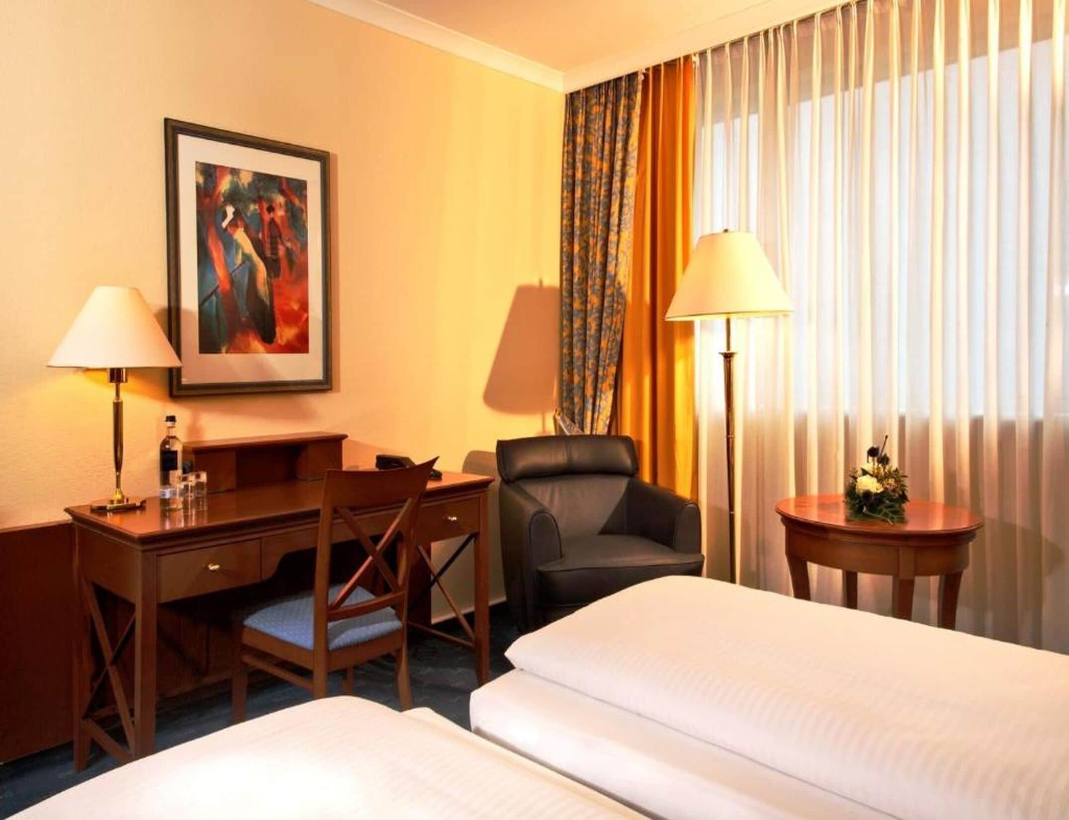 A hotel room with two beds, a desk with a chair, a desk lamp, a chair, a framed picture on the wall, and a window with curtains.