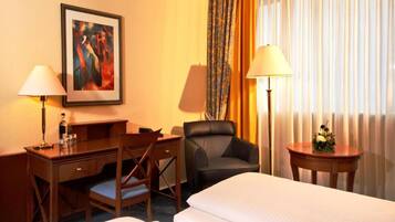 A hotel room with two beds, a desk with a chair, a desk lamp, a chair, a framed picture on the wall, and a window with curtains.