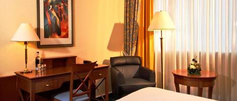 Une chambre d’hôtel avec deux lits, un bureau avec une chaise, une lampe de bureau, une chaise, un tableau encadré au mur et une fenêtre avec des rideaux.