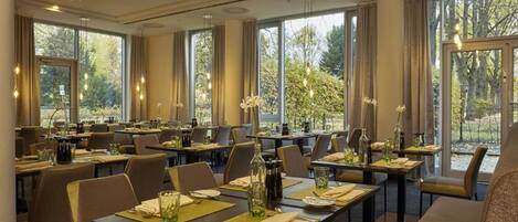 A dining area with tables set for service, large windows with curtains, and a view of trees.