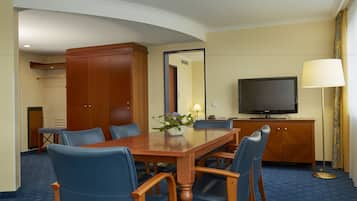 A hotel room with a wooden dining table, blue upholstered chairs, a television, and a large mirror.