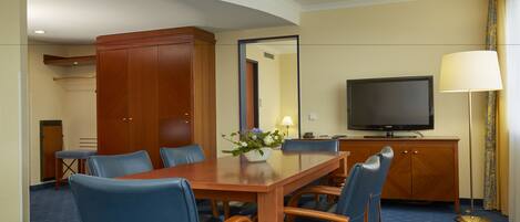 A hotel room with a wooden dining table, blue upholstered chairs, a television, and a large mirror.
