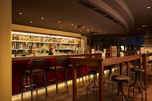 Zona de bar con varios taburetes, una barra repleta de botellas y una persona sirviendo.