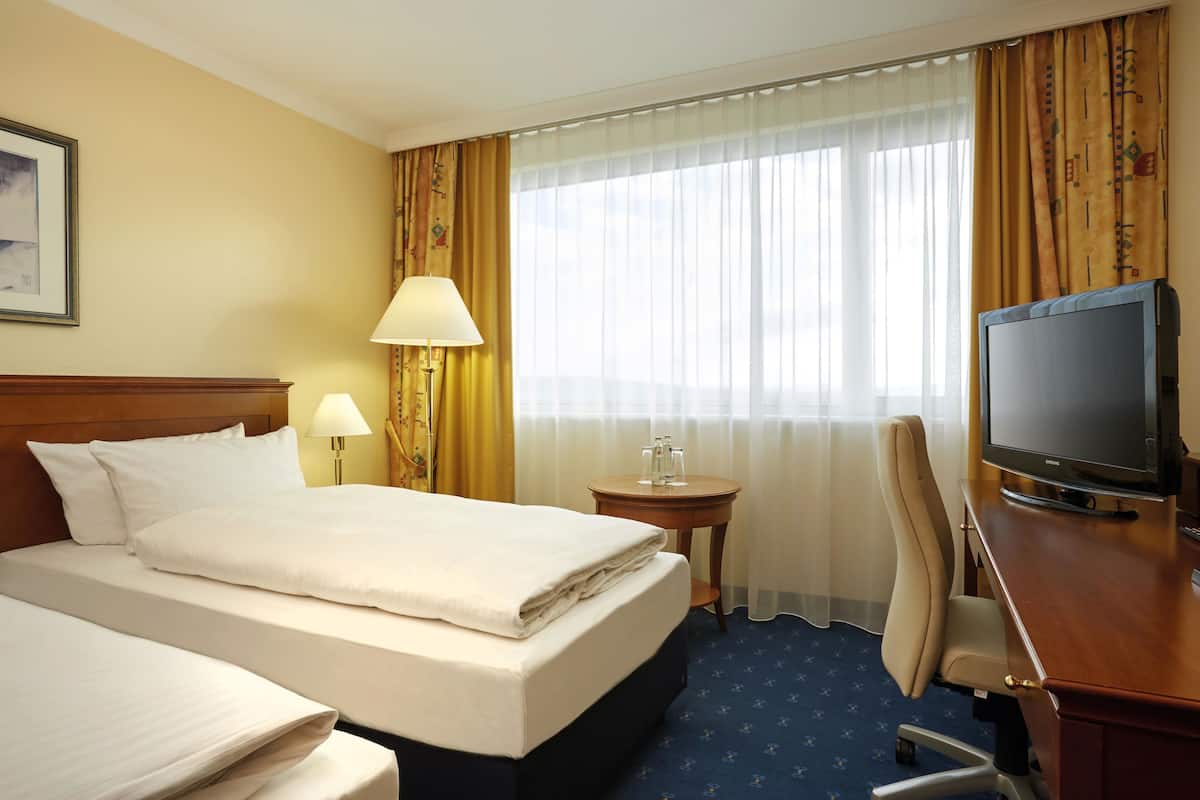 A hotel room with two beds, a desk with a television, a lamp, and a small table with glasses.