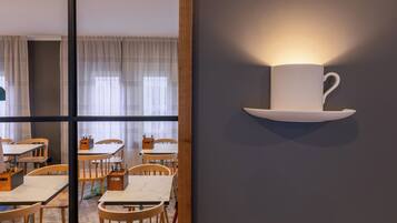 A modern dining area with wooden tables and chairs, a large window with sheer curtains, and a wall-mounted light fixture shaped like a cup.