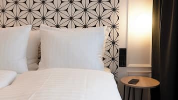 Una cama bien tendida con sábanas y almohadas blancas, una mesita de madera redonda con un teléfono inteligente, y una pared con un diseño geométrico.