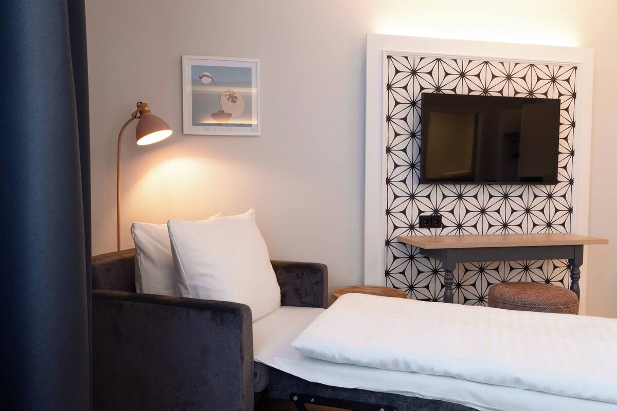 Ein modernes Zimmer mit einem Bett, einem Schreibtisch, einem Sessel, einer Lampe und einem an einer Wand mit geometrischem Muster befestigten Fernseher.