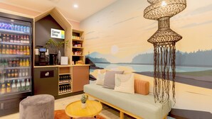 A cozy lounge area with a sofa, ottomans, a coffee table, a beverage dispenser, and a mural of a sunset landscape. - Garner Hotel Mannheim City by IHG (Mannheim)