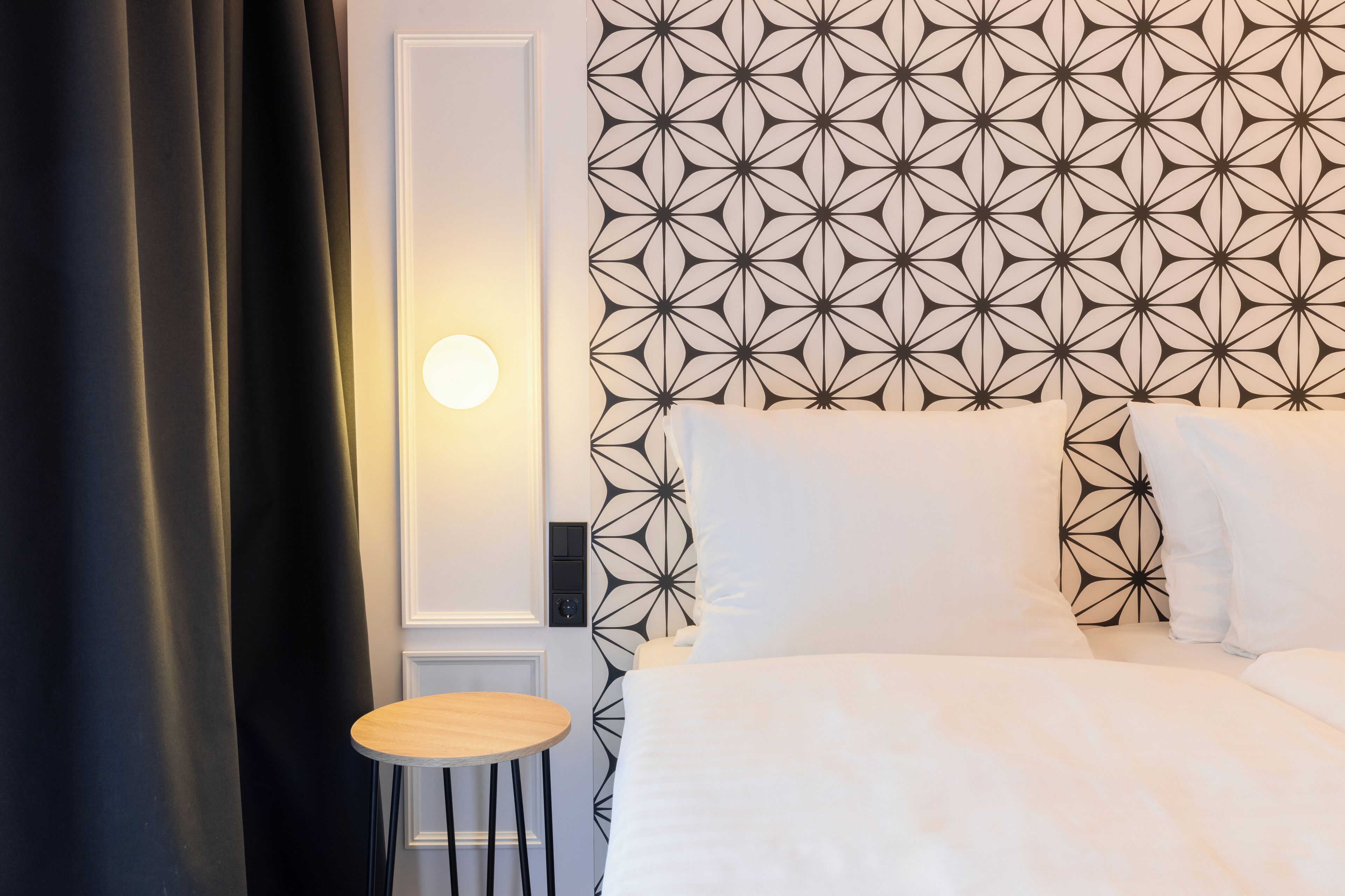 Una habitación de hotel con un papel tapiz de diseño geométrico, una mesa redonda de madera y una cama con ropa blanca.