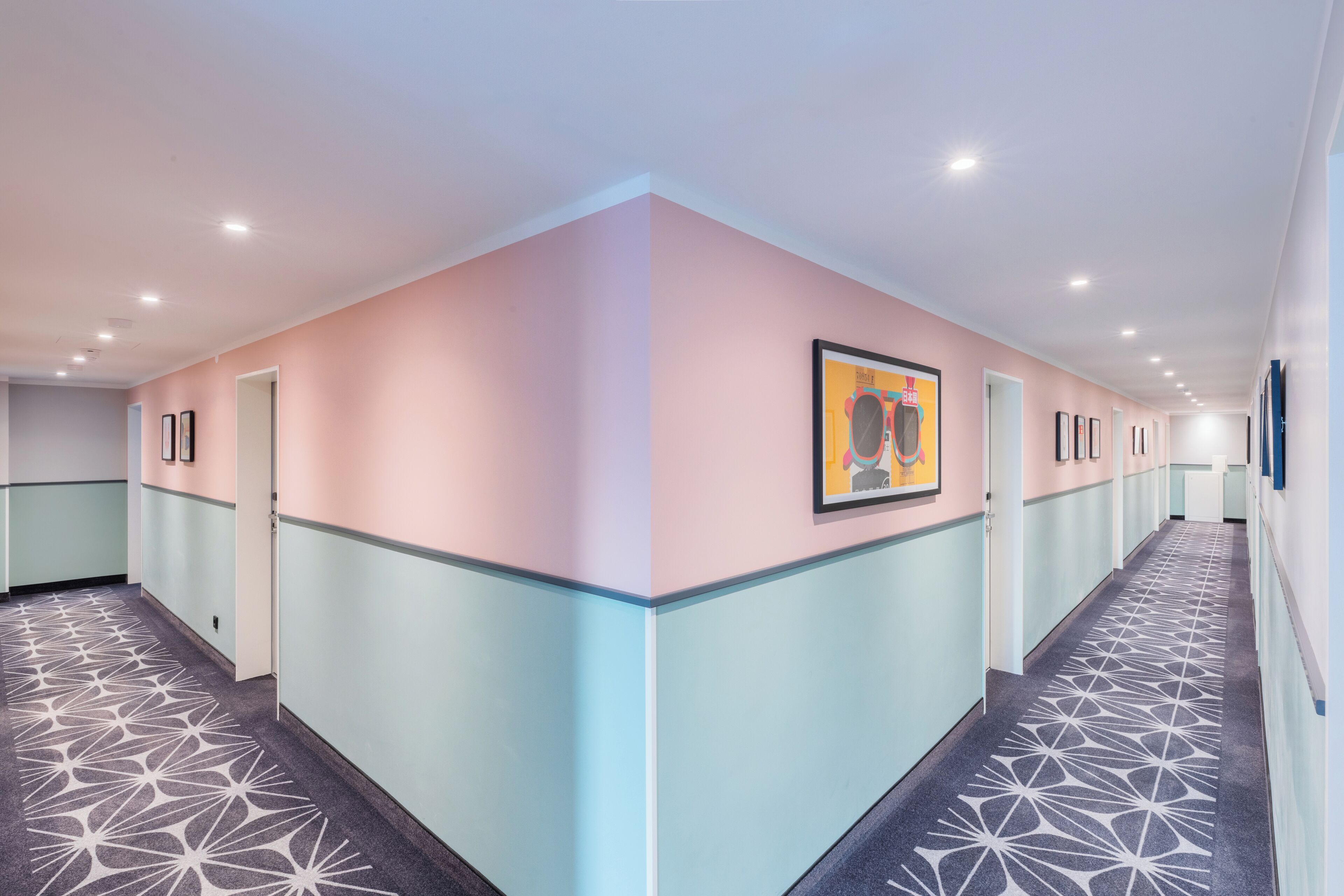 A hotel corridor with a geometric patterned carpet, pastel-colored walls, and framed artwork on the walls.