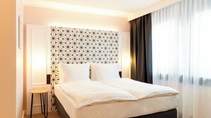 A neatly made bed with white linens, a geometric-patterned headboard, a small wooden stool, and a view of a cityscape through sheer curtains. - Garner Hotel Mannheim City by IHG (Mannheim)