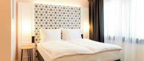 A neatly made bed with white linens, a geometric-patterned headboard, a small wooden stool, and a view of a cityscape through sheer curtains.