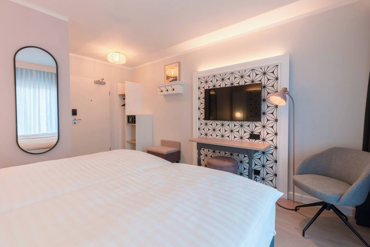 Ein modernes Hotelzimmer mit einem großen Bett, einem Schreibtisch, einem Flachbildfernseher, einem Sessel und einem Spiegel.