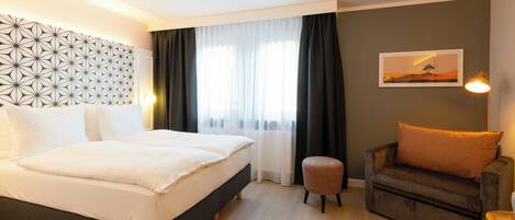 Quarto de hotel com cama, sofá, um banquinho pequeno, luminária na parede e um quadro.